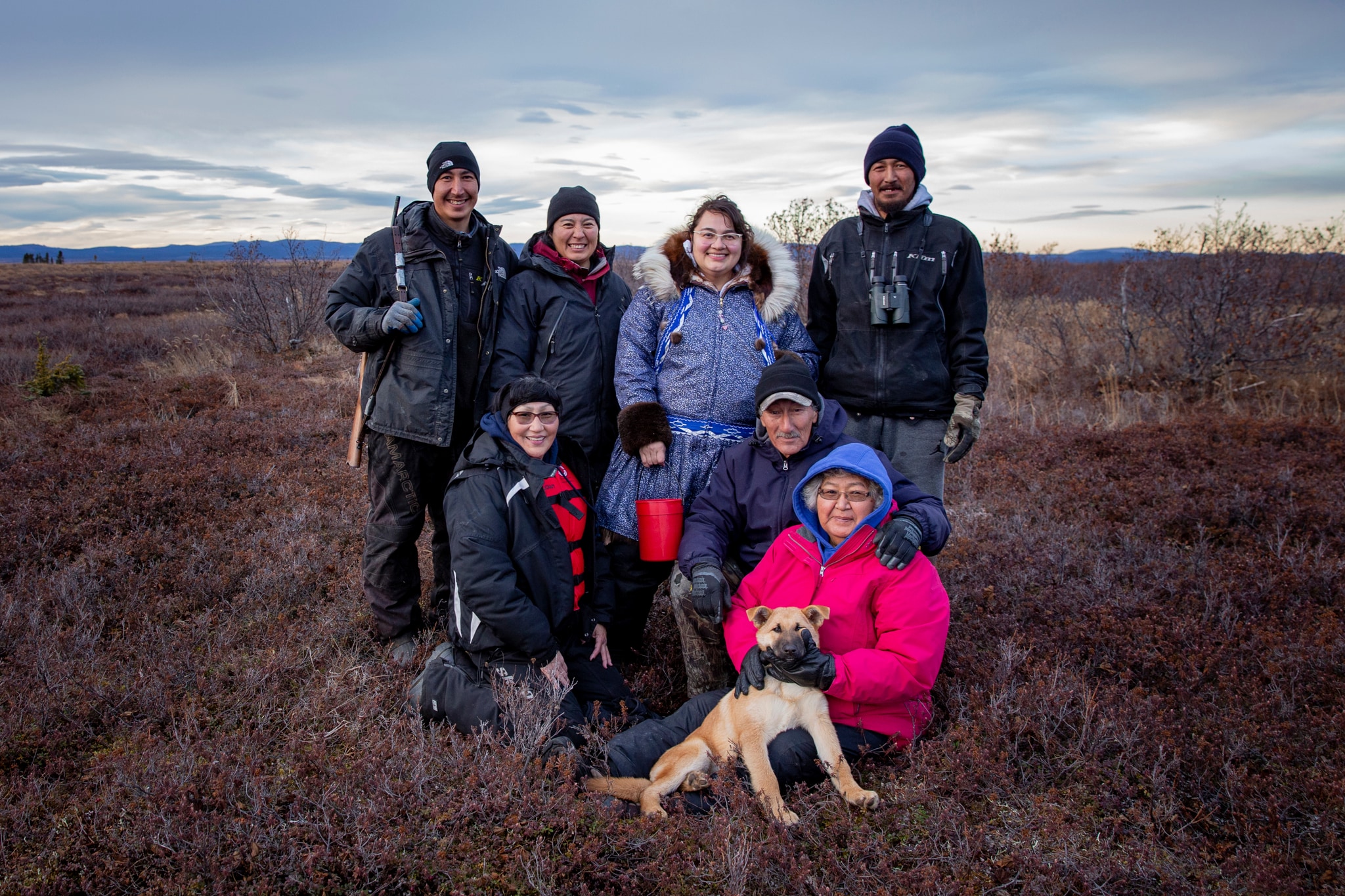 A family of seven smiling people and their dog standing outside.