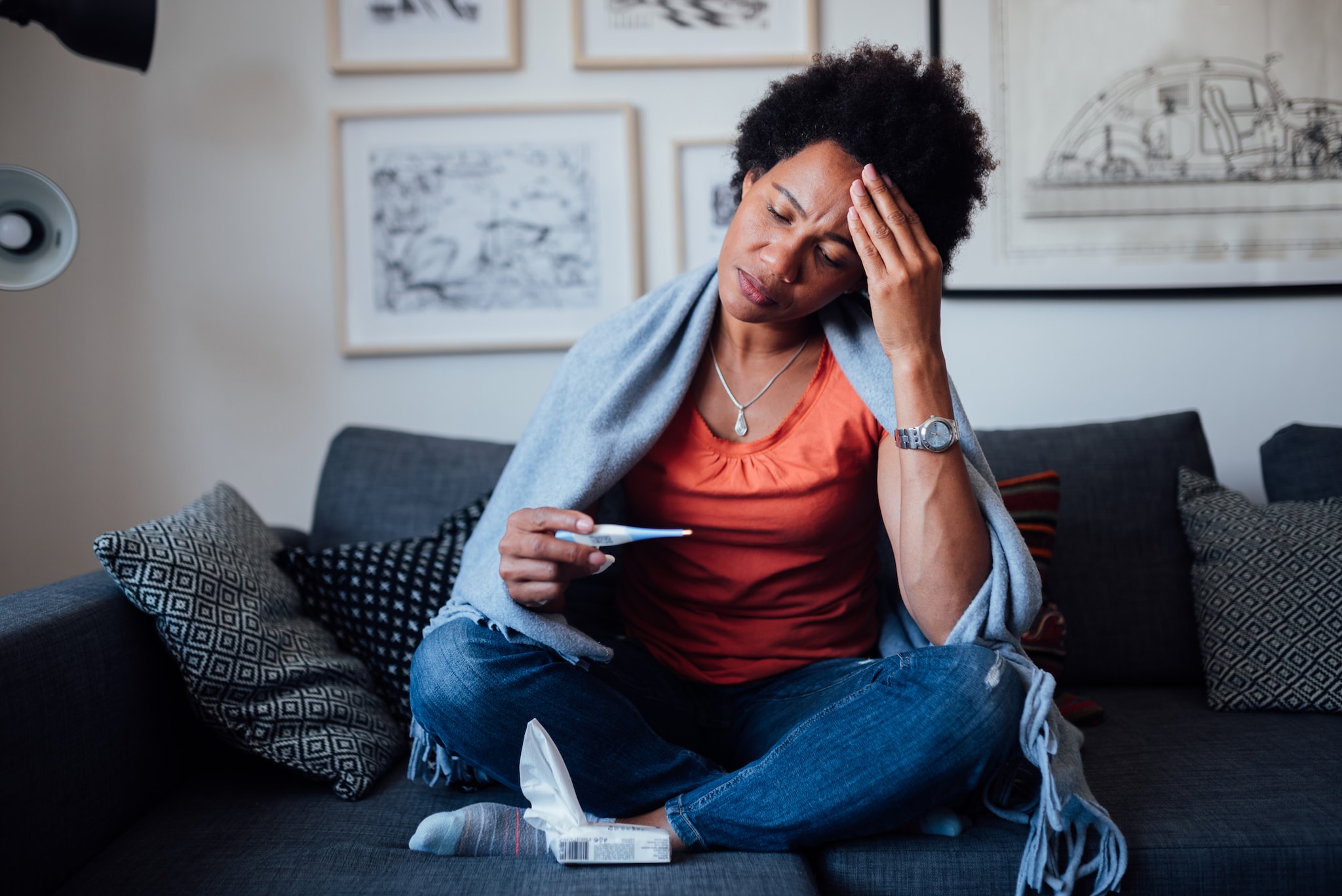 A woman sitting on a sofa holding her head and checking the thermometer