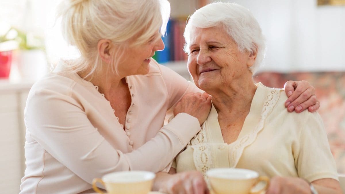 Older woman caregiver with white hair having tea with a much older woman with white hair.