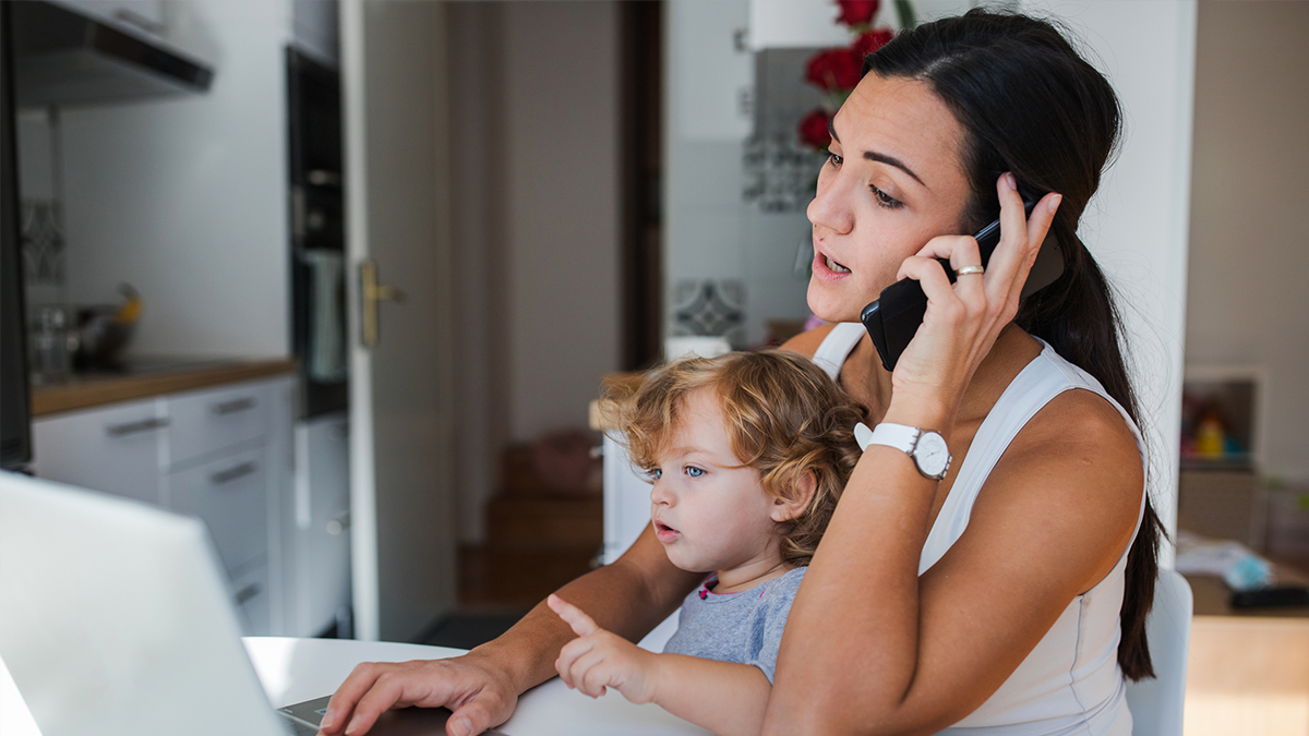 A mother is on the phone while holding a baby and looking at the computer