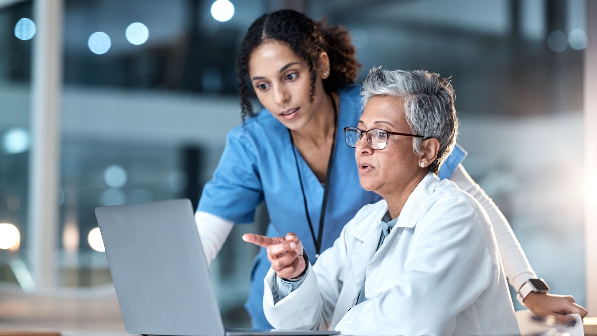 Two healthcare workers talking to each other while looking at a laptop. One worker is pointing at the laptop.