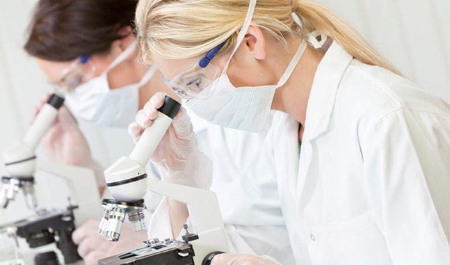 Dos técnicos de laboratorio con batas blancas, mascarillas y gafas de protección mirando a través de microscopios.