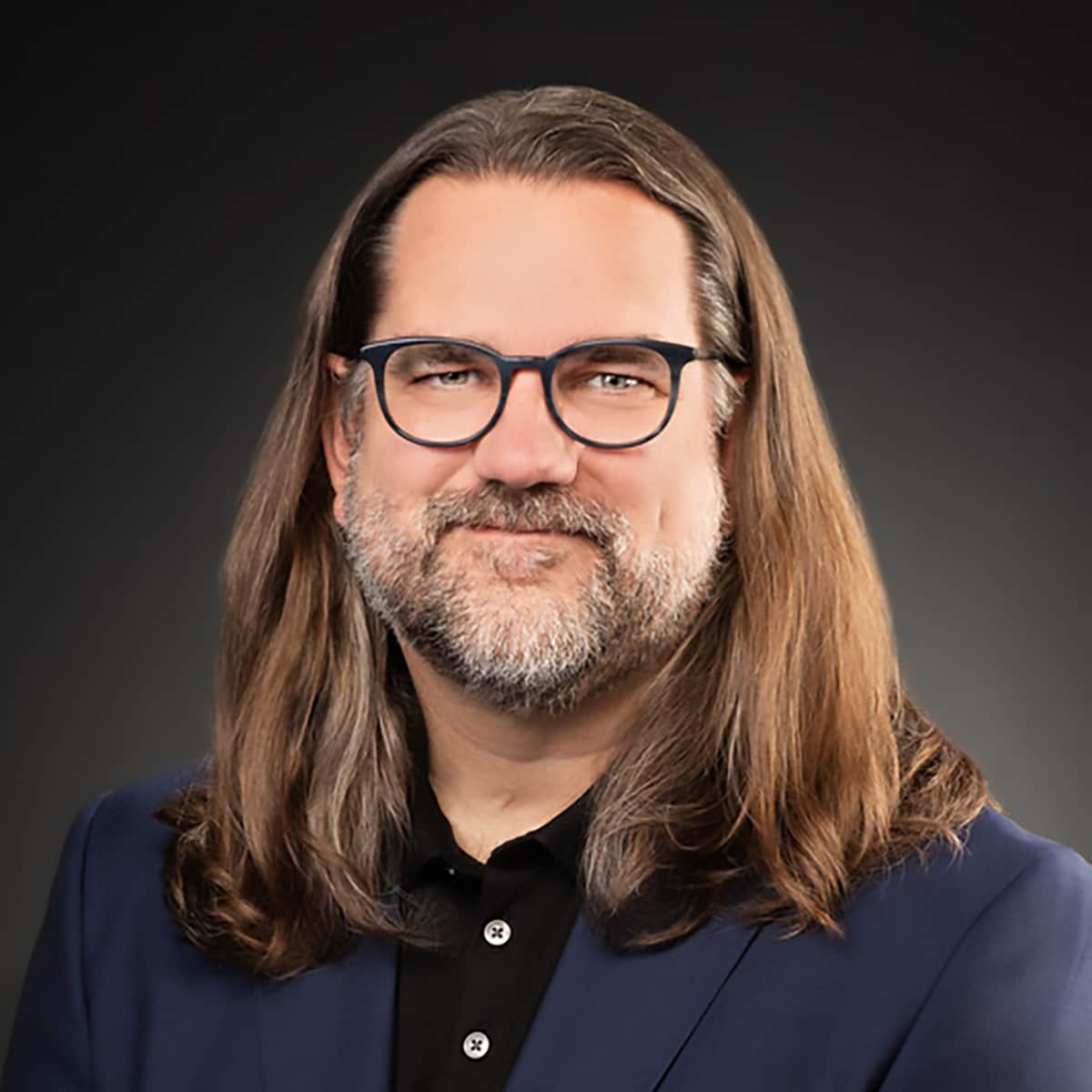 Dylan George, Ph.D., has long brown hair and short brown beard, wearing a dark blue shirt against a grey background.