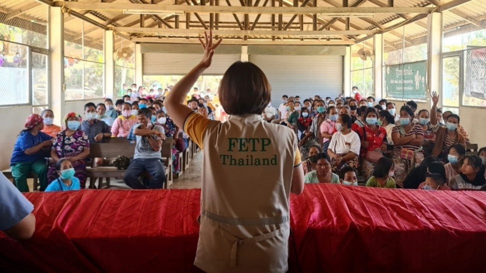 Asian woman in a tan vest hand signaling to an audience.