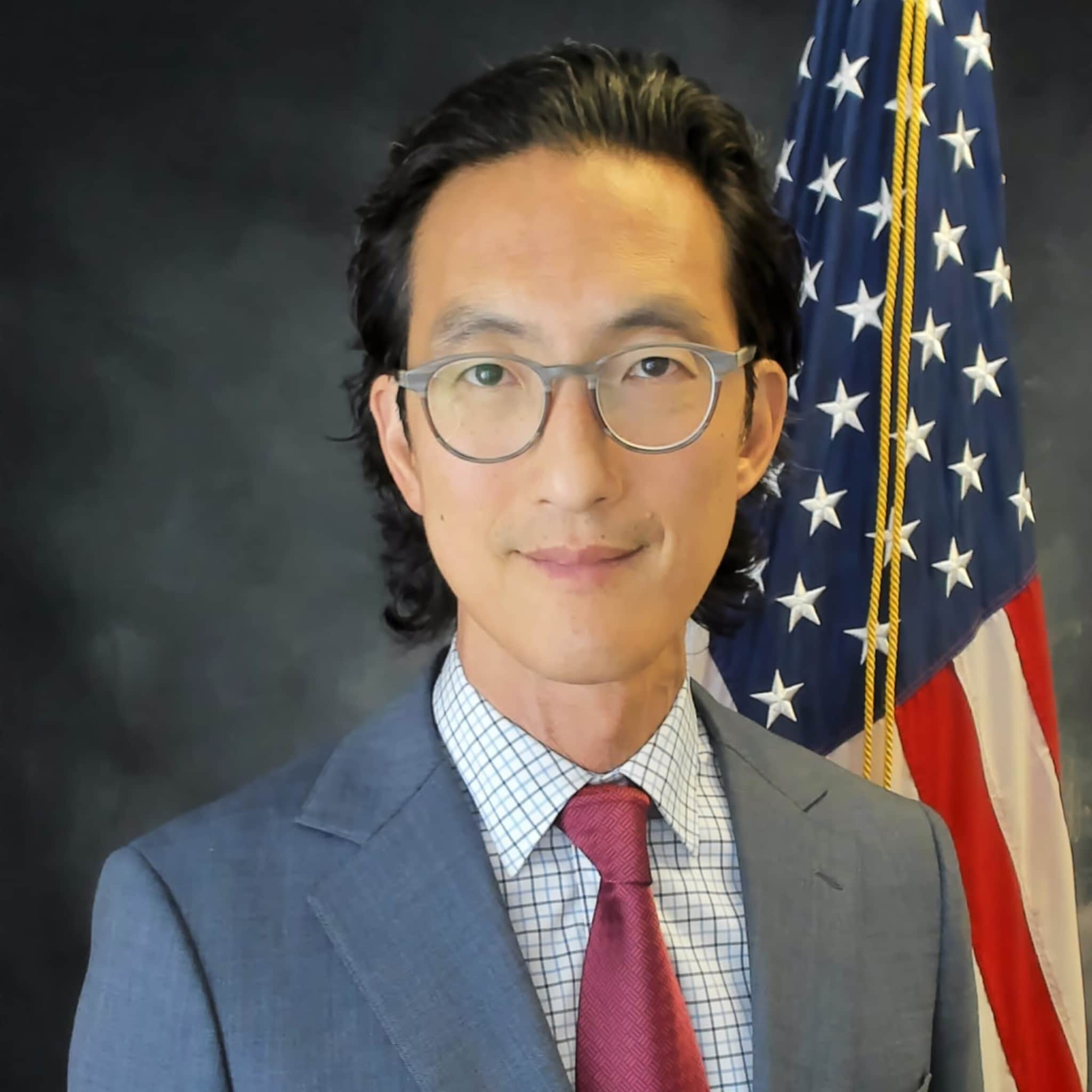 A man with black hair, dark eyes, and glasses. He is wearing a grey suit jacket, checkered shirt, and burgundy tie.