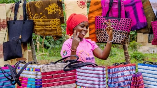 A young woman in pink stands at with bags she created.