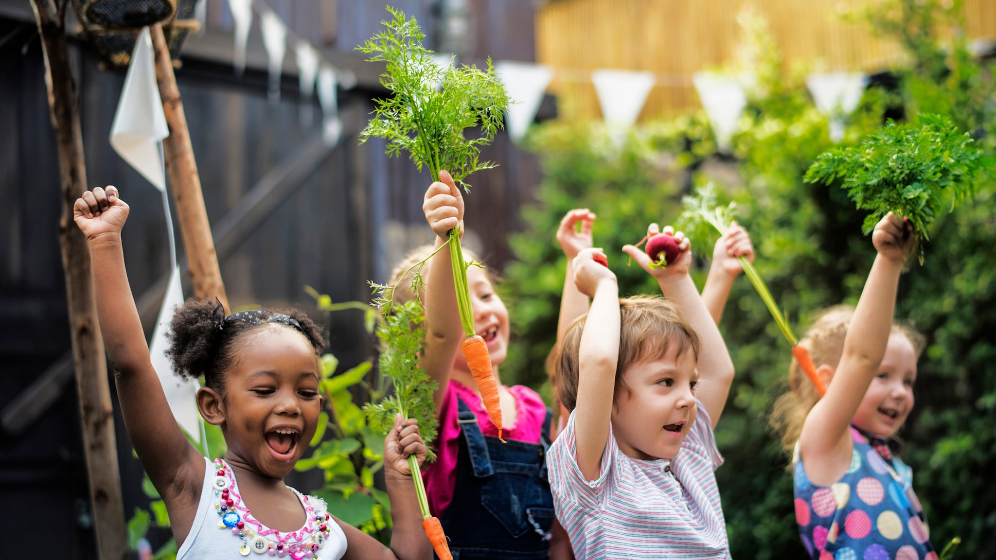 Children in a garden, with their hands up in the air, holding picked greens.