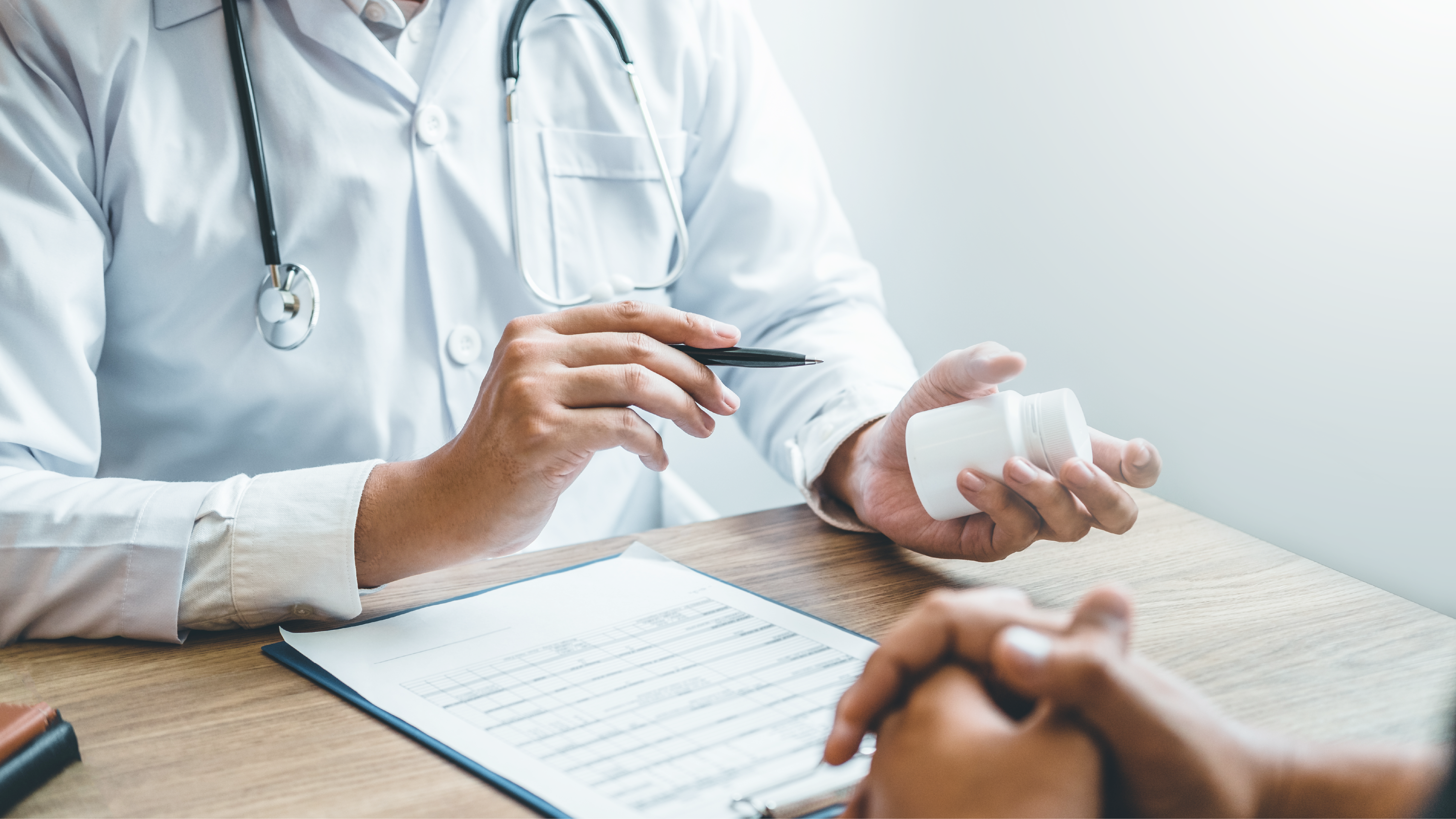 A health care provider’s hands and body, sitting at a table facing a patient, holding and pointing to a pill bottle.