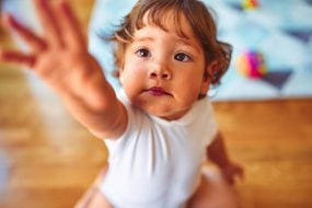 Toddler boy sitting on the floor and reaching up to the camera.