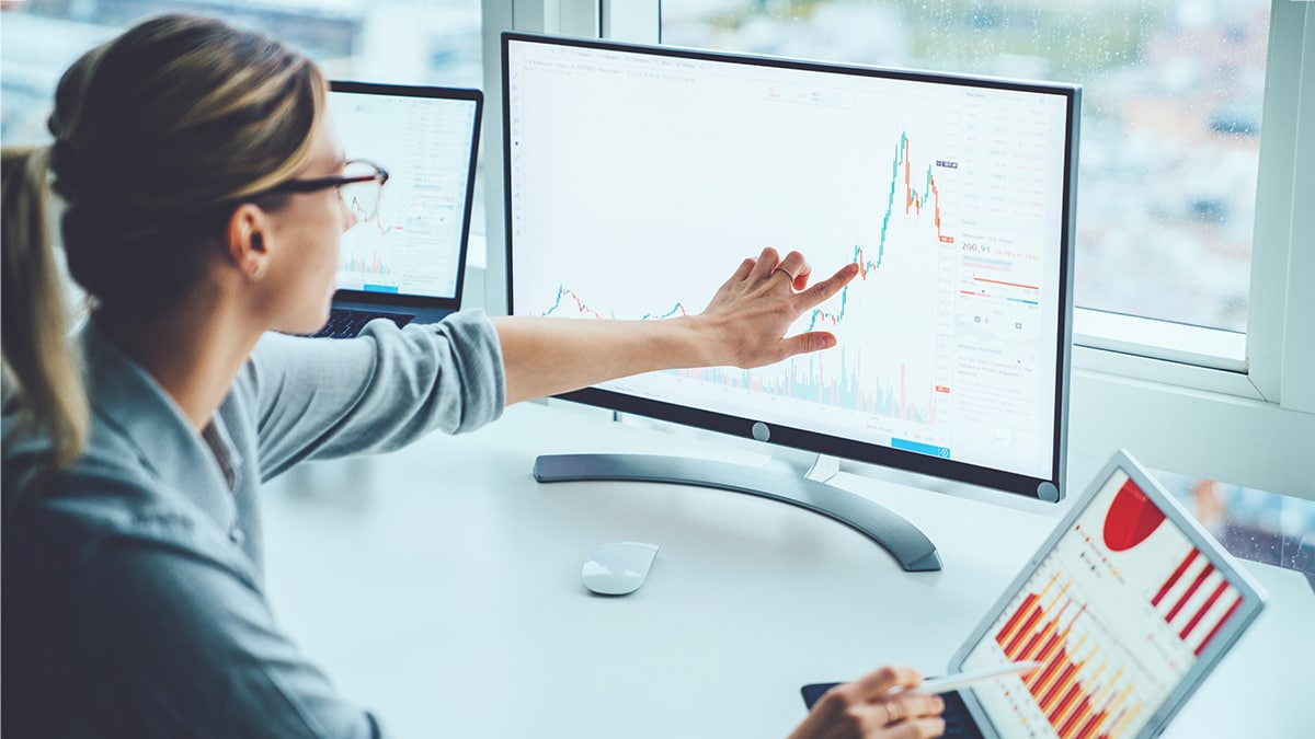 Woman in glasses compares line graph on a computer monitor to bar graph on a laptop screen.