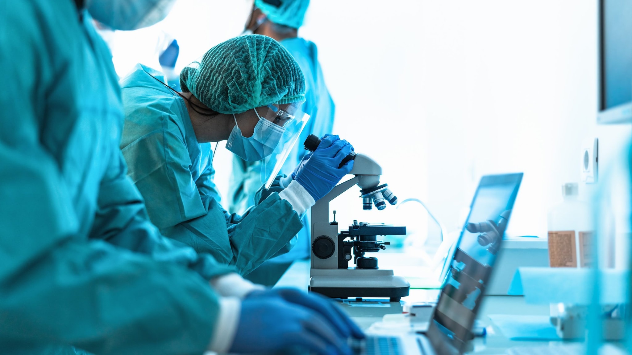 person wearing protective glasses and clothing looking into a microscope