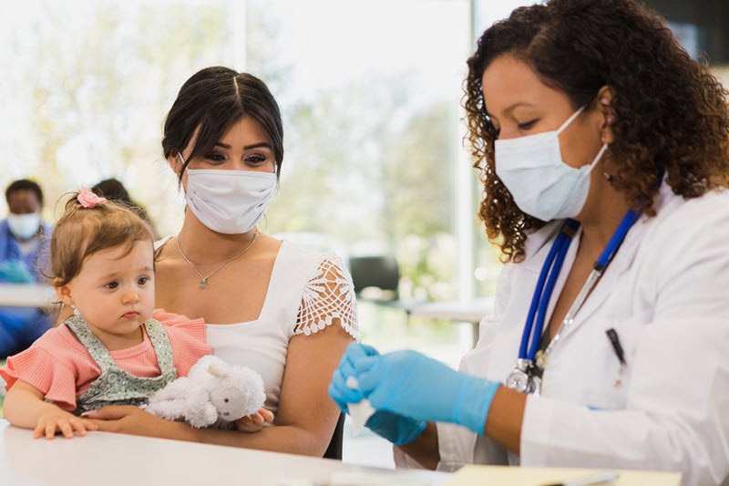 A mother holds her baby while a health care provider prepares to give the baby a vaccination.