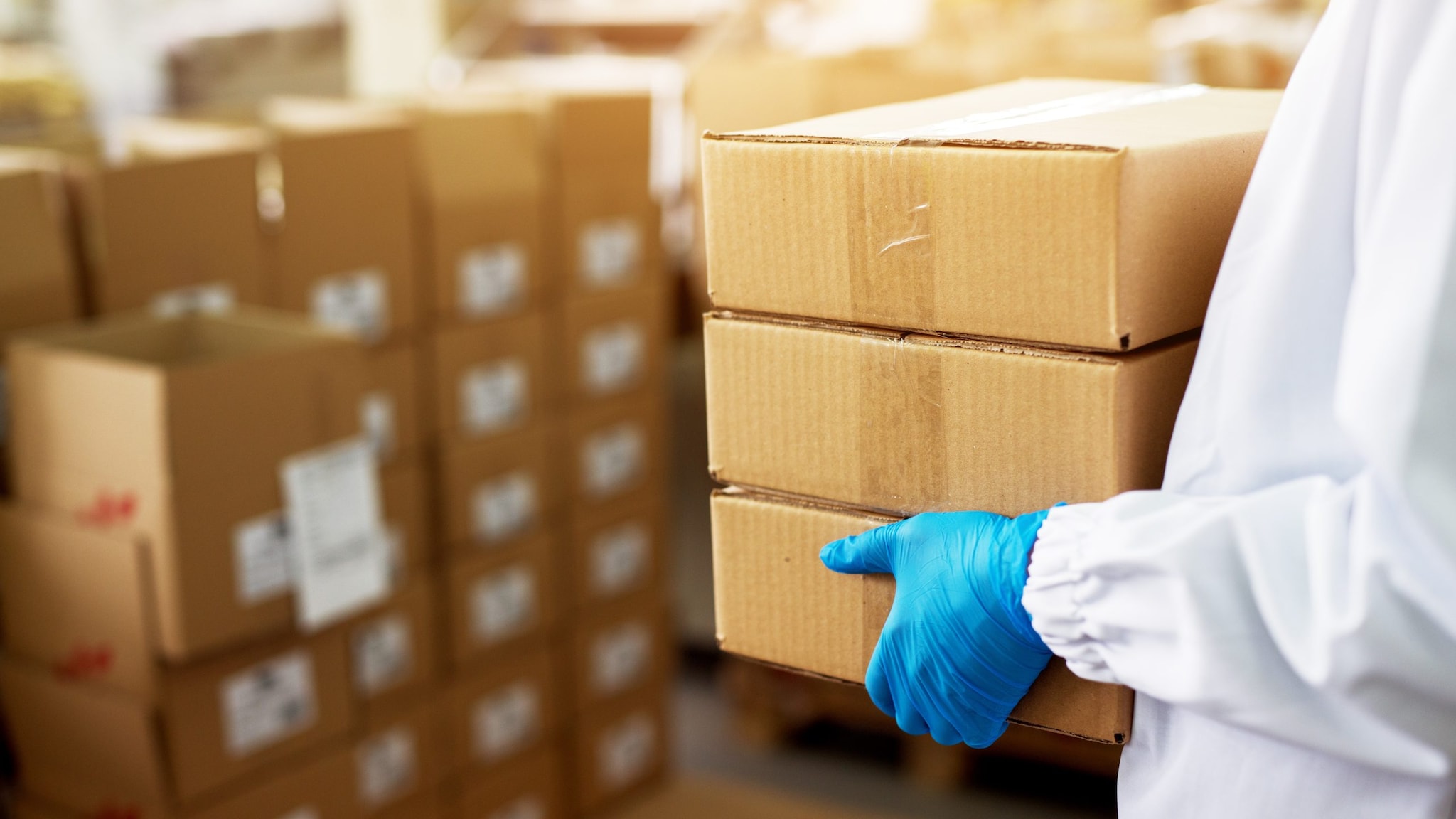 Person wearing a lab coat and blue gloves carrying cardboard boxes.