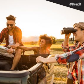 Image displays a man reading a map while sitting in the back of a pick-up truck, with a woman ostensibly pointing toward their destination, and another man looking in that direction through a pair of binoculars.