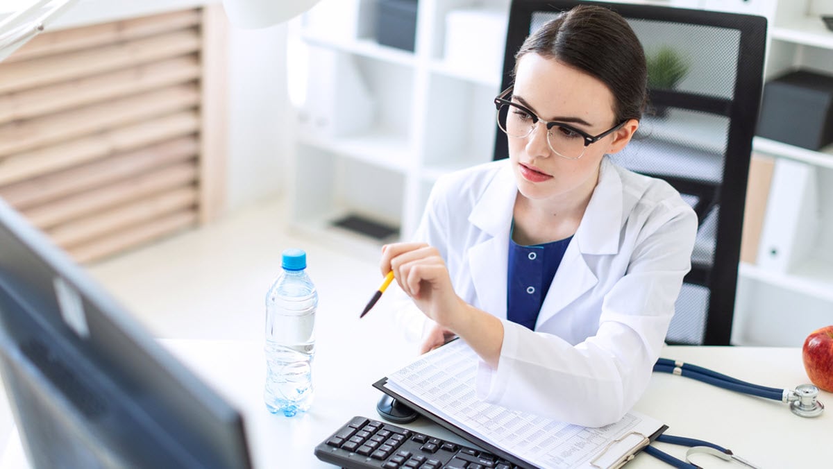 A female health care provider is sitting at a computer desk with documents and a pen in her hands.