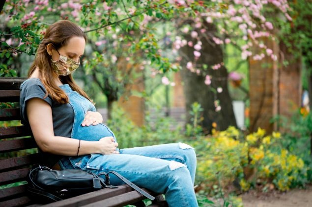 A pregnant woman wearing a cloth facemask sits on a park bench holding her belly.