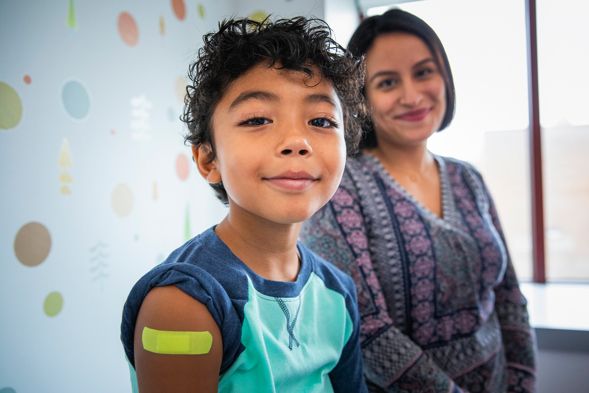 Pediatric patient with band-aid on upper arm smiling in medical office with parent smiling in the background.