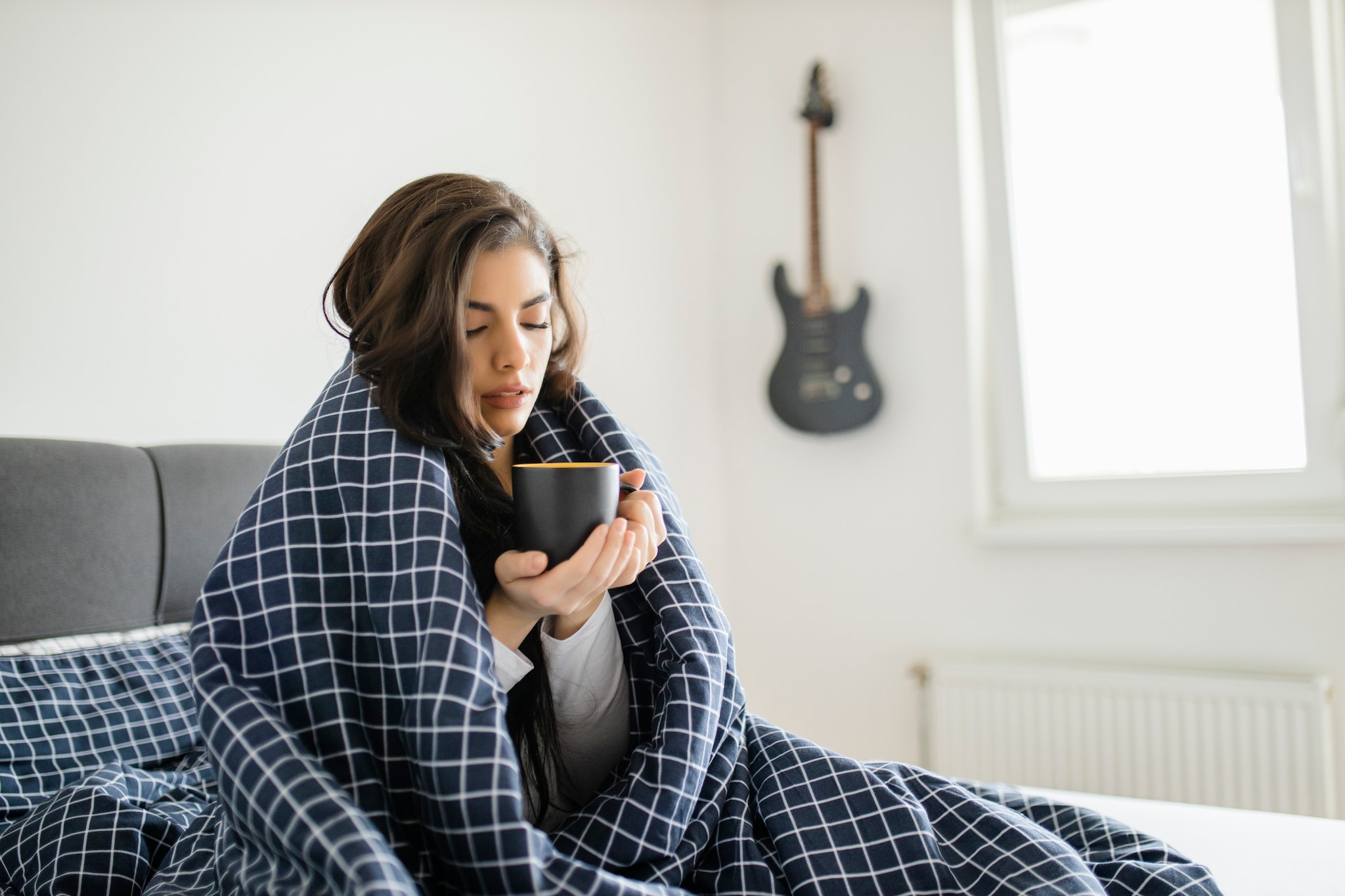 A woman sitting in bed covered in a blanket and holding a cup in her hands.