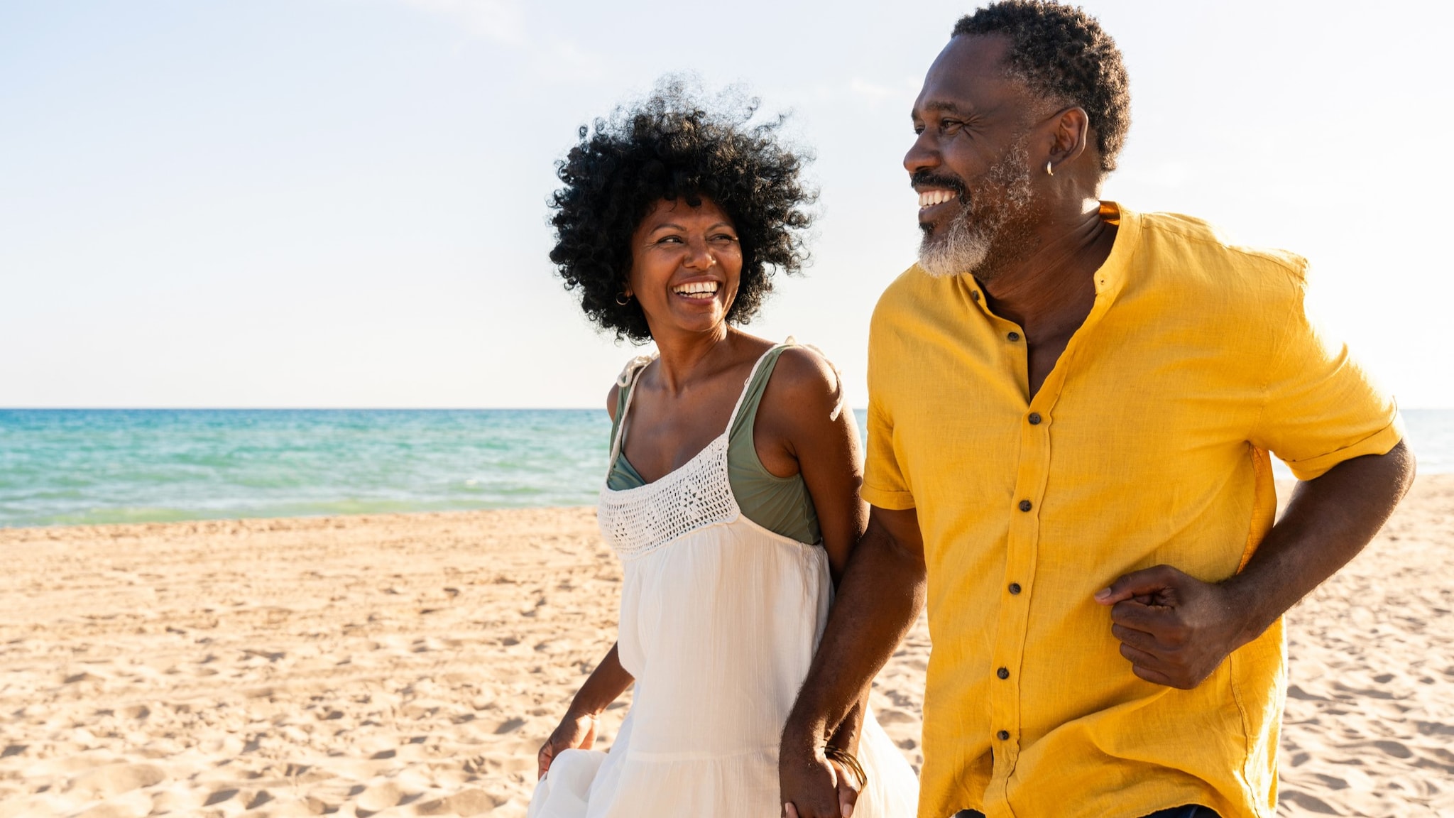 An older couple holding hands on the beach, walking on the sand.