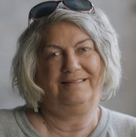 White female wearing a gray shirt with gray hair with sunglasses sitting on top of her head.